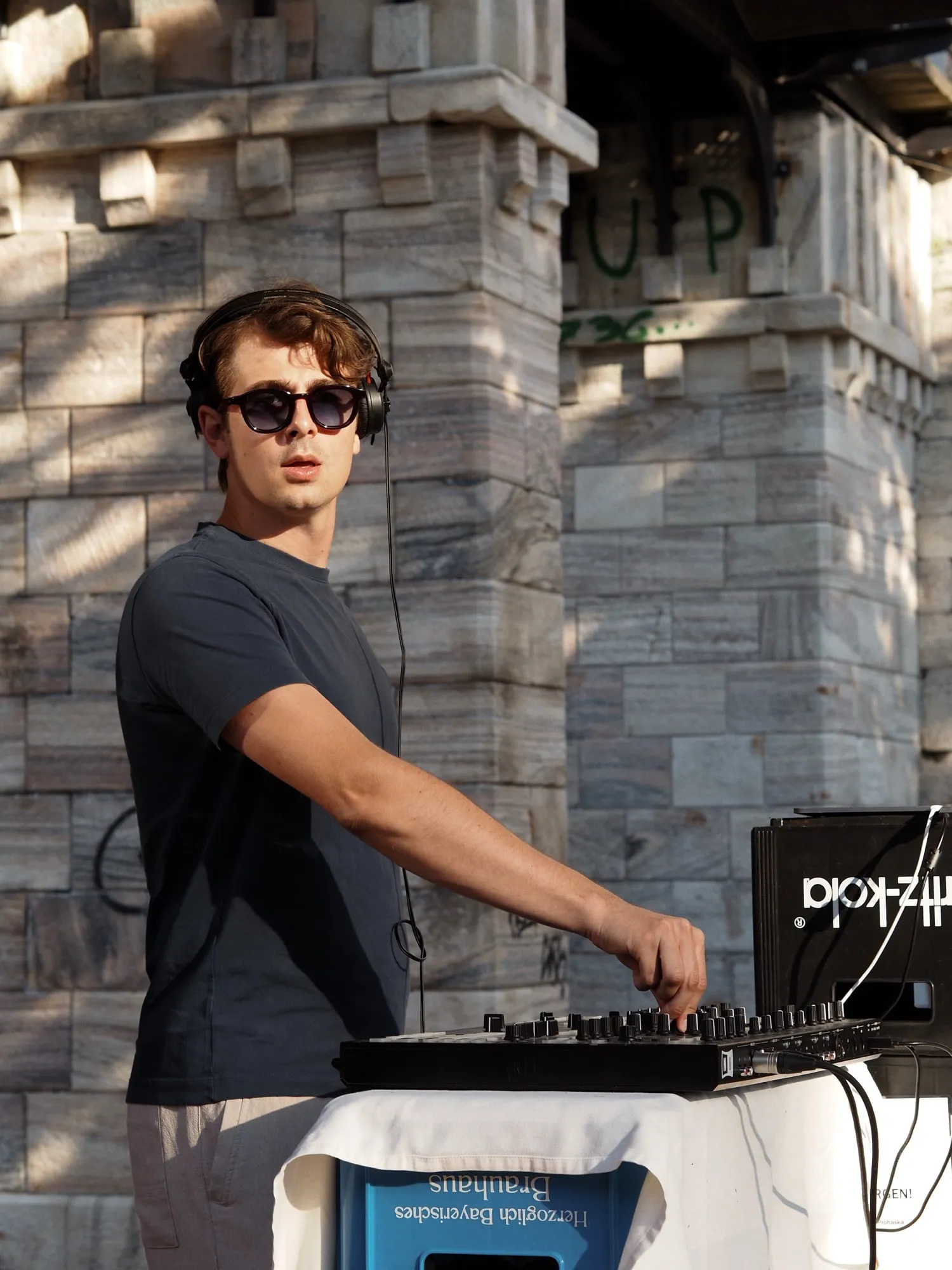 Plie behind the decks at an outdoor event, a stone wall behind him